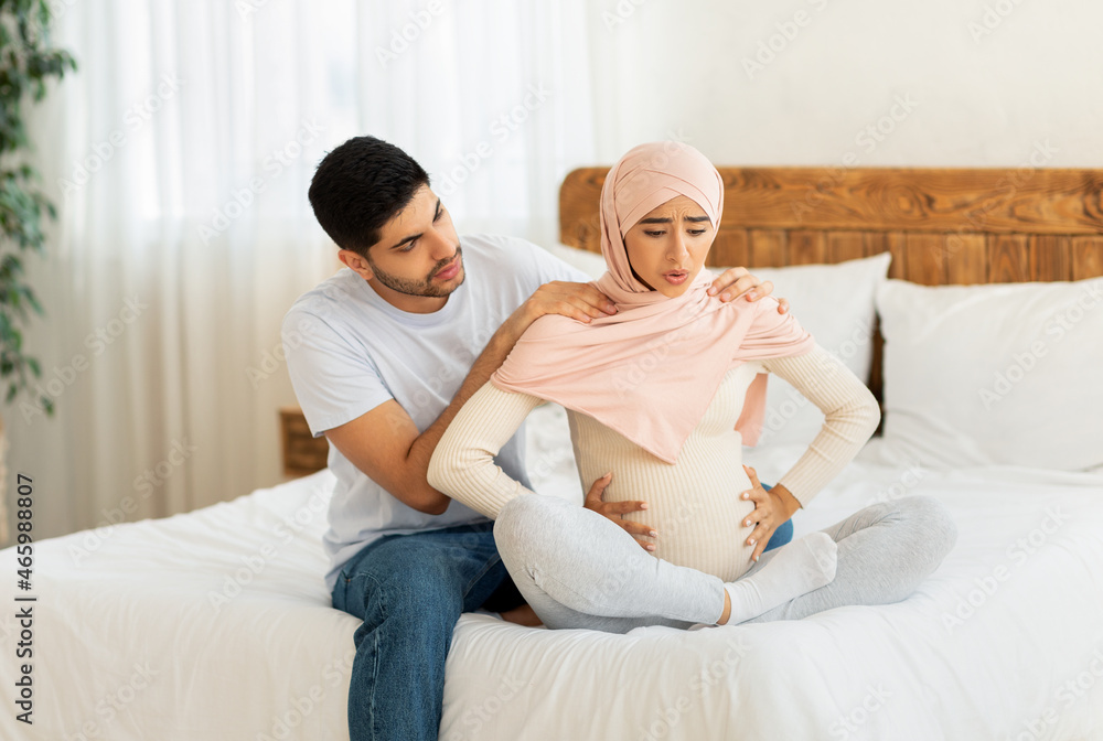 Pregnant muslim woman having labor pains, sitting by husband on bed, arab man massaging wife's shoulders Pregnant muslim woman having labor pains, sitting by husband on bed, arab man massaging wife's shoulders