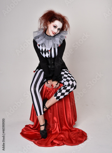 Full length  portrait of red haired  girl wearing a black and white clown jester costume  theatrical circus character.  Sitting down on chair  isolated on  studio background.