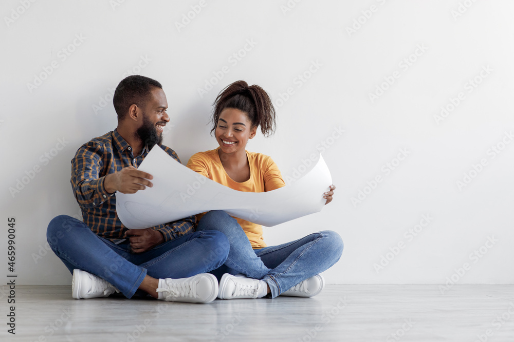 Happy funny young african american guy and lady look at blueprints at home, sit on floor over white wall background Happy funny young african american guy and lady look at blueprints at home, sit on floor over white wall background