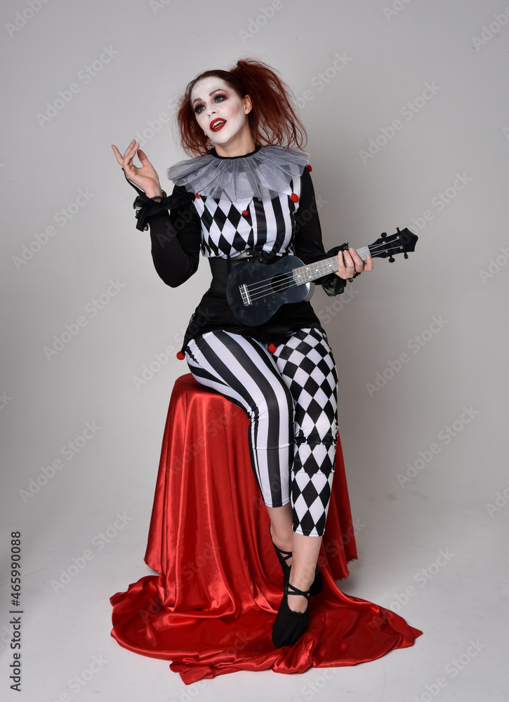 Full length portrait of red haired girl wearing a black and white clown jester costume, theatrical circus character. Sitting down on chair, isolated on studio background. Full length portrait of red haired girl wearing a black and white clown jester costume, theatrical circus character. Sitting down on chair, isolated on studio background.