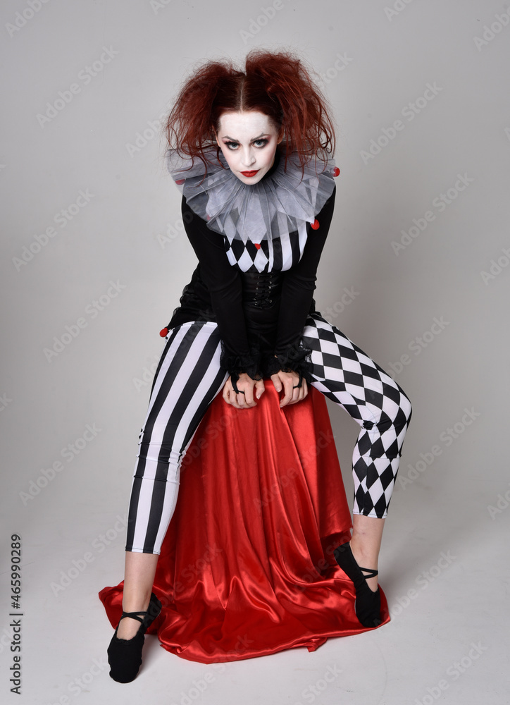 Full length portrait of red haired girl wearing a black and white clown jester costume, theatrical circus character. Sitting down on chair, isolated on studio background. Full length portrait of red haired girl wearing a black and white clown jester costume, theatrical circus character. Sitting down on chair, isolated on studio background.