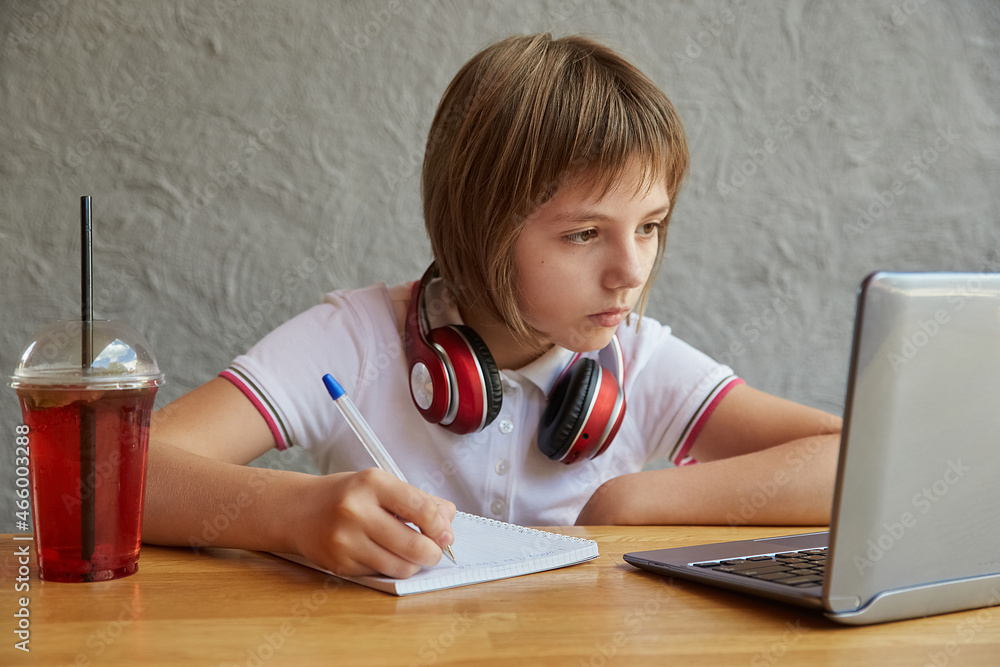 schoolgirl with headphones sits at table, watching at laptop screen, writing notes at copybook. little caucasian girl studying with computer, doing homework. distant learning, online education, lesson schoolgirl with headphones sits at table, watching at laptop screen, writing notes at copybook. little caucasian girl studying with computer, doing homework. distant learning, online education, lesson