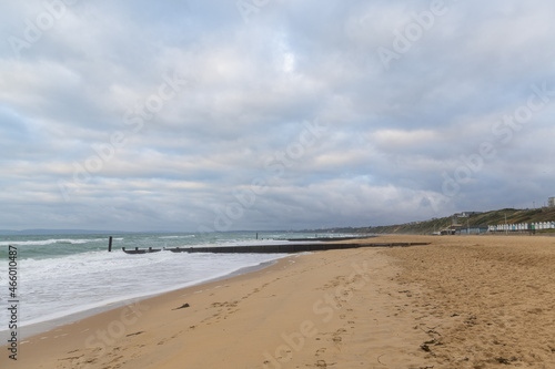 A scenic view of a sandy beach with crashing waves  wooden groynes and some grassy dunes in the background under a grey stormy sky