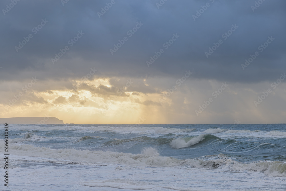 A scenic view of a raging sea with huge crashing waves with a majestc ray of light coming through a cloudy stormy sky with some hill in the background