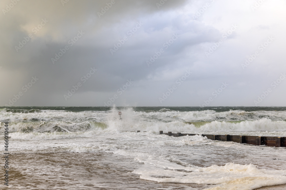 A dramatic view of a very choppy sea with huge crashing waves on groynes (breakwater) during a major storm under a grey sky