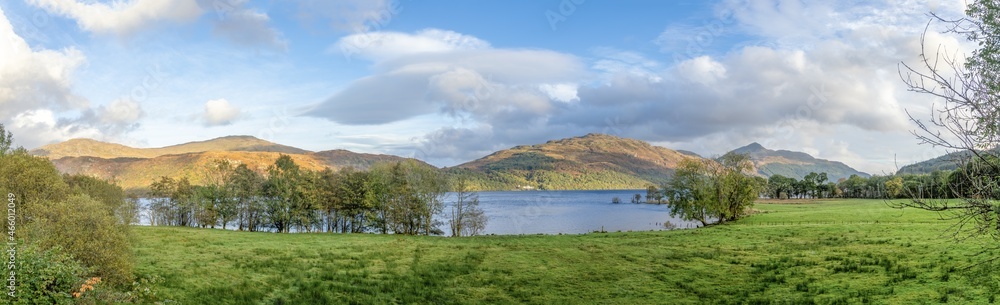 A scenic panoramic view of a Scottish lake with majestic colorful mountain range in the backgound under a  beautiful blue sky and some white clouds