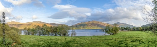 A scenic panoramic view of a Scottish lake with majestic colorful mountain range in the backgound under a beautiful blue sky and some white clouds