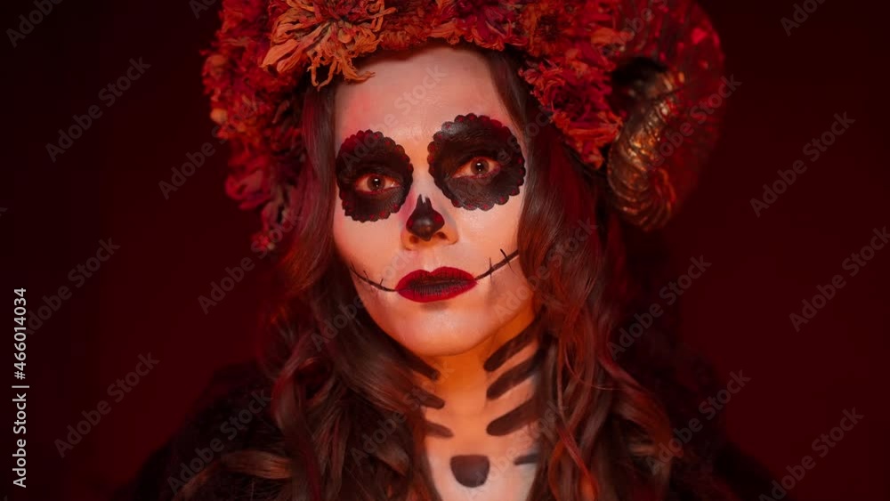 Caucasian female brunette in a Halloween image of a witch with the makeup of a painted skull with ram horns, sewn her mouth, flowers looking into the camera in a dark fiery room close-up Caucasian female brunette in a Halloween image of a witch with the makeup of a painted skull with ram horns, sewn her mouth, flowers looking into the camera in a dark fiery room close-up