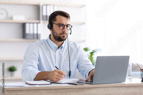 Serious mature guy in glasses and headphones works at computer, support clients, watches webinar or online lesson