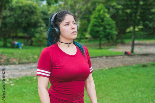 beautiful college woman with blue hair and red dress, listening to music with her wireless headphones in a park, relaxed girl thinking about her future while walking in the park. concept of relaxation
