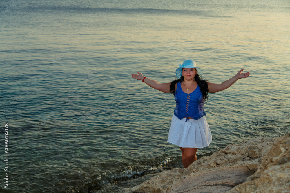 A young woman in a white skirt and a sun hat stands on the shore against the backdrop of the sea at sunset with her arms outstretched.