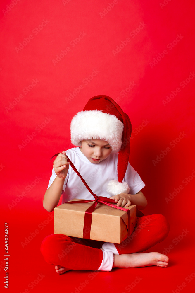 Merry Christmas. Portrait of a cute boy in a santa cap with gift boxes on a red background. A place for text. Merry Christmas. Portrait of a cute boy in a santa cap with gift boxes on a red background. A place for text.