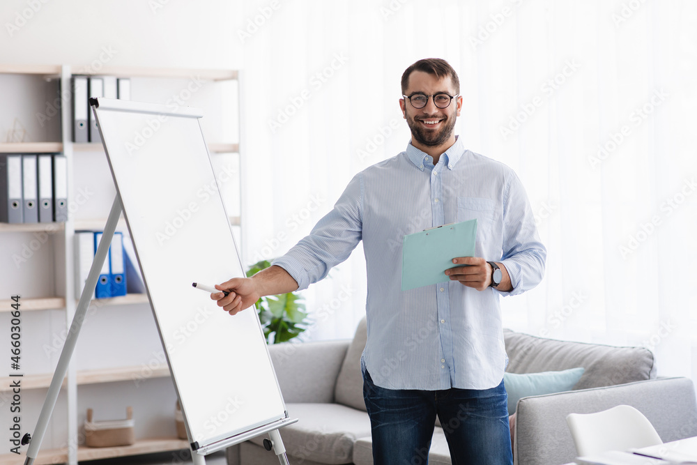 Friendly happy adult european male teacher with beard in glasses shows on blackboard teaches students