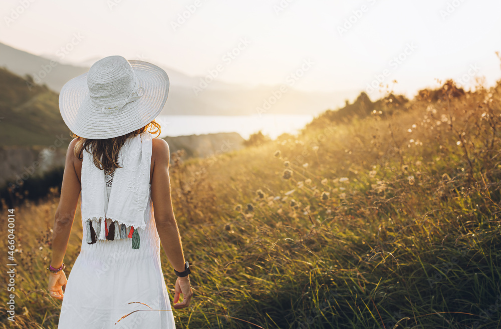 a woman with her back turned in a white dress, looks towards a sunset. she is in a beautiful meadow with golden natural light.