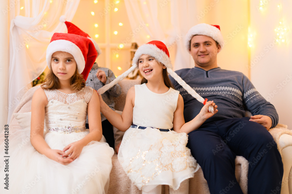 Fototapeta premium family portrait - parent and children in home interior decorated with holiday lights and gifts, dressed in santa hat for new year celebration