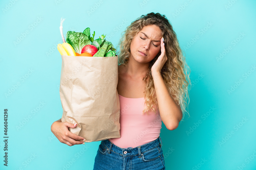 Girl with curly hair holding a grocery shopping bag isolated on green background with headache Girl with curly hair holding a grocery shopping bag isolated on green background with headache