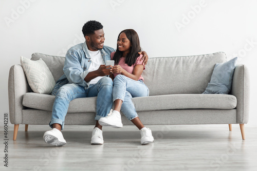 Happy black couple sitting on couch, sharing using cellphone