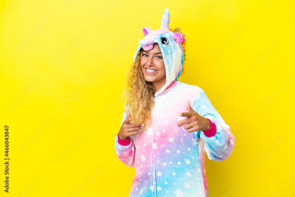 Girl with curly hair wearing a unicorn pajama isolated on yellow background pointing to the front and smiling
