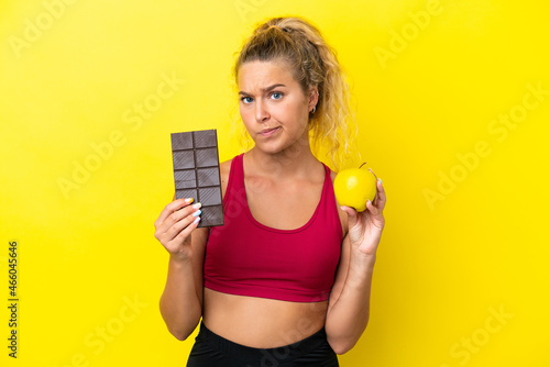 Girl with curly hair isolated on yellow background taking a chocolate tablet in one hand and an apple in the other