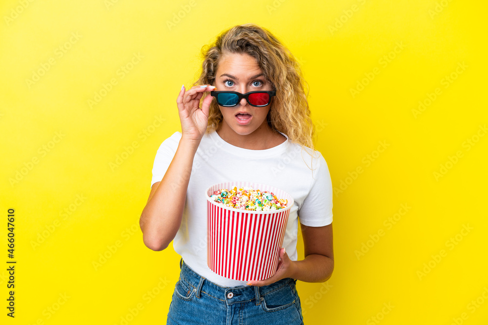 Obraz premium Girl with curly hair isolated on yellow background surprised with 3d glasses and holding a big bucket of popcorns
