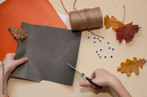 a child cuts out a Halloween craft from black paper against a beige background, toy eyes, jute rope, and dry leaves