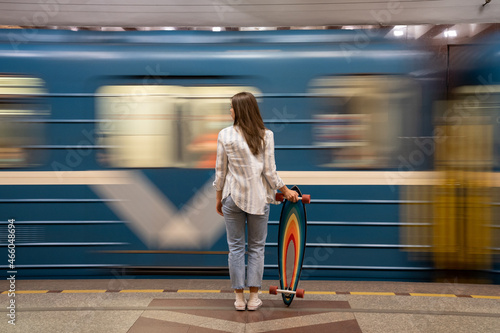 Fotomurale Young girl passenger with longboard standing on subway station platform with blurry moving blue train on background, rear view