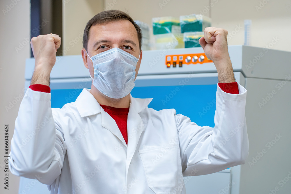 Happy excited scientist or doctor man wearing white coat celebrating success with arms raised in room with laboratory box in UV light for sterilization. Successful male of scientific laboratory Happy excited scientist or doctor man wearing white coat celebrating success with arms raised in room with laboratory box in UV light for sterilization. Successful male of scientific laboratory
