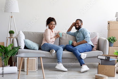 Smiling young african american man and woman relax on couch, drinking coffee in room with cardboard boxes