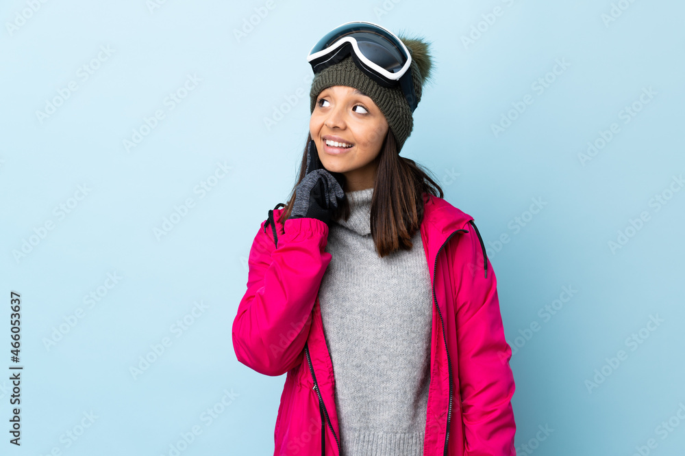 Mixed race skier girl with snowboarding glasses over isolated blue background thinking an idea while looking up.