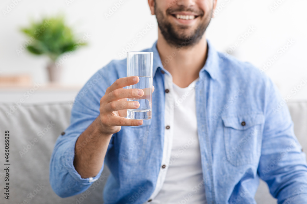 Smiling middle aged caucasian guy with beard sits on sofa and holds glass with clean still water Smiling middle aged caucasian guy with beard sits on sofa and holds glass with clean still water