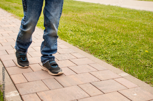 A child in blue jeans and dark sneakers with blue inserts is walking on a sidewalk tile against a background of green grass