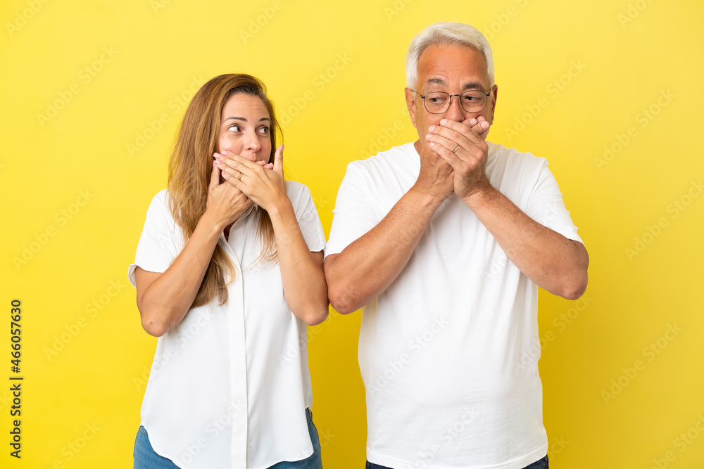 Middle age couple isolated on yellow background covering mouth with hands Middle age couple isolated on yellow background covering mouth with hands