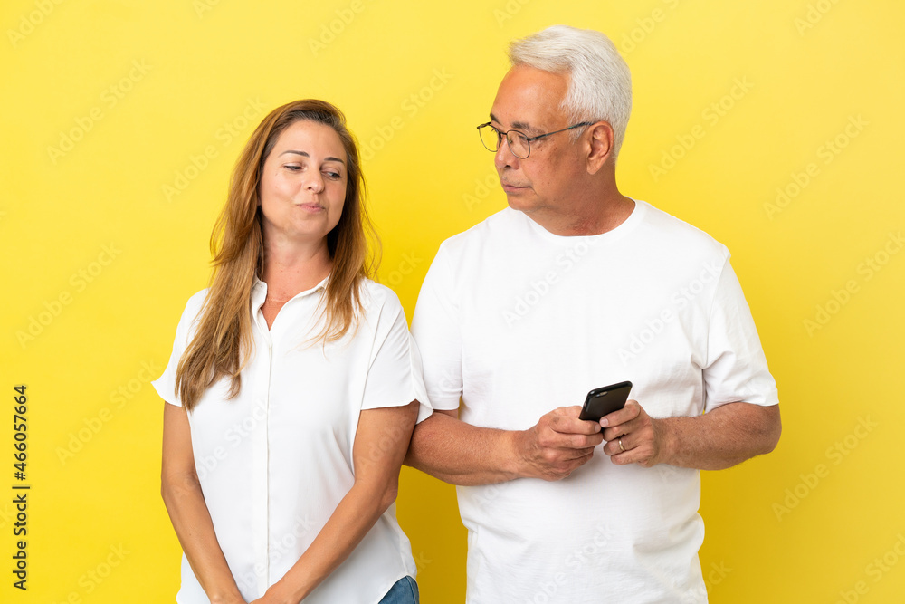 Middle age couple isolated on yellow background reading a message with the mobile Middle age couple isolated on yellow background reading a message with the mobile