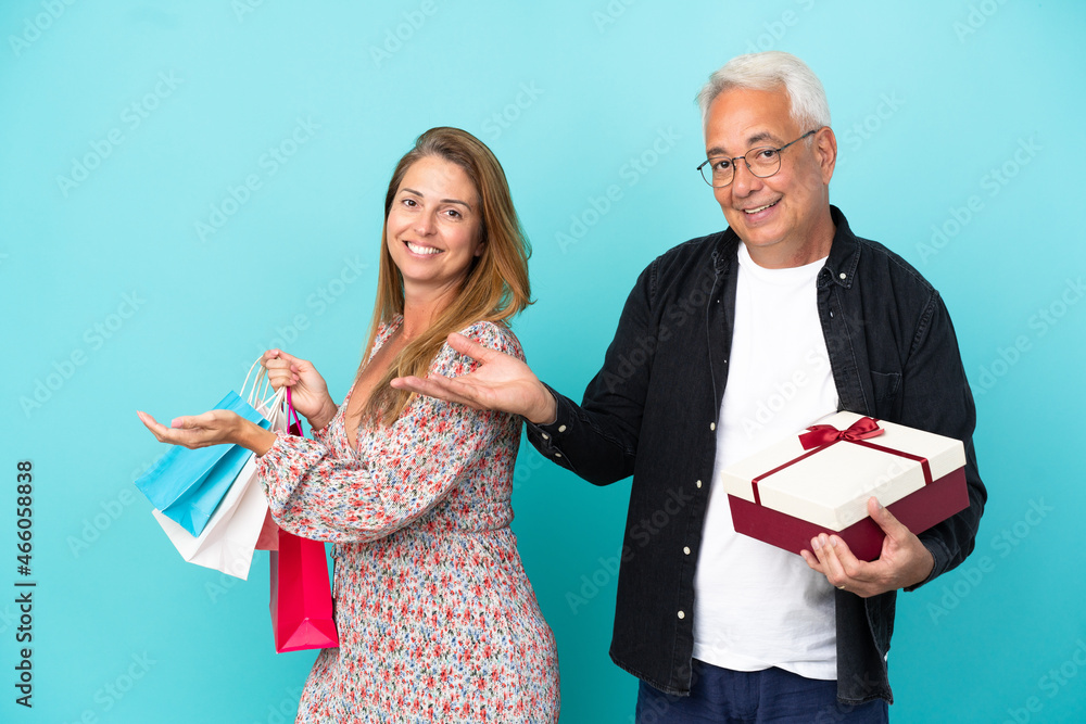 Middle age couple with shopping bag and gift isolated on blue background extending hands to the side for inviting to come Middle age couple with shopping bag and gift isolated on blue background extending hands to the side for inviting to come