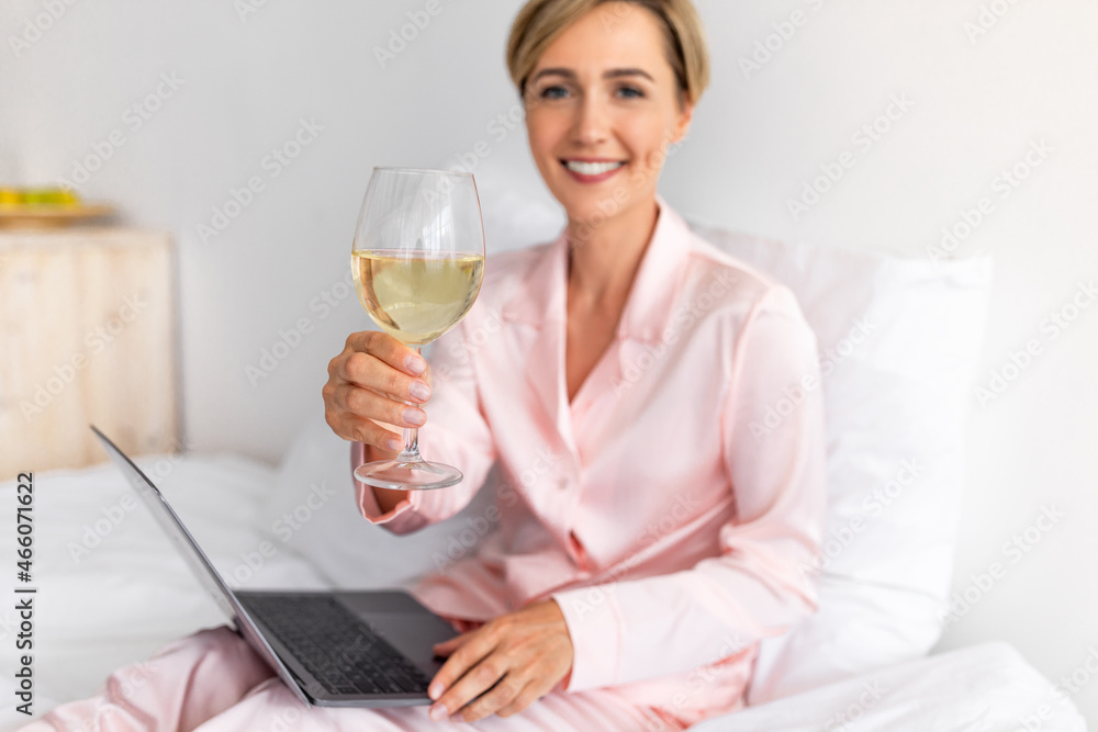 Happy woman sitting on bed using laptop drinking white wine Happy woman sitting on bed using laptop drinking white wine