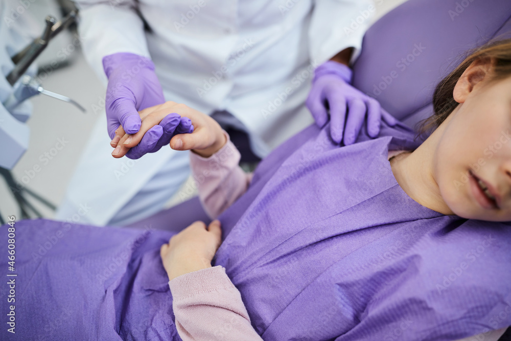 Close-up of scared girl holds dentist's hand during dental procedure at dentist's office.
