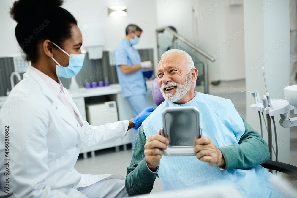 Satisfied senior man looks his teeth in a mirror and talks with his dentist at dental clinic. Satisfied senior man looks his teeth in a mirror and talks with his dentist at dental clinic.