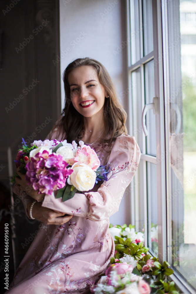 Beautiful young woman with brown hair and red lips, dressed in a pink dress sits on the windowsill. Portrait of happy smiling girl holds a bouquet of different flowers in her hands. Spring mood. Beautiful young woman with brown hair and red lips, dressed in a pink dress sits on the windowsill. Portrait of happy smiling girl holds a bouquet of different flowers in her hands. Spring mood.