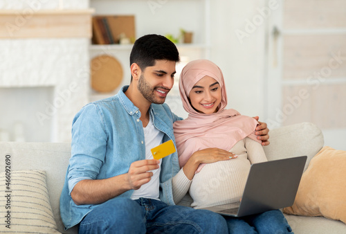 Preparing for childbirth. Loving muslim pregnant couple using laptop computer and credit card, sitting on sofa