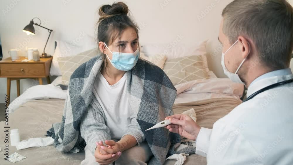 Doctor measuring temperature of young woman in medical mask with thermometer. Male doctor checking temperature to young woman during home visit. Coronavirus protection during the quarantine Doctor measuring temperature of young woman in medical mask with thermometer. Male doctor checking temperature to young woman during home visit. Coronavirus protection during the quarantine