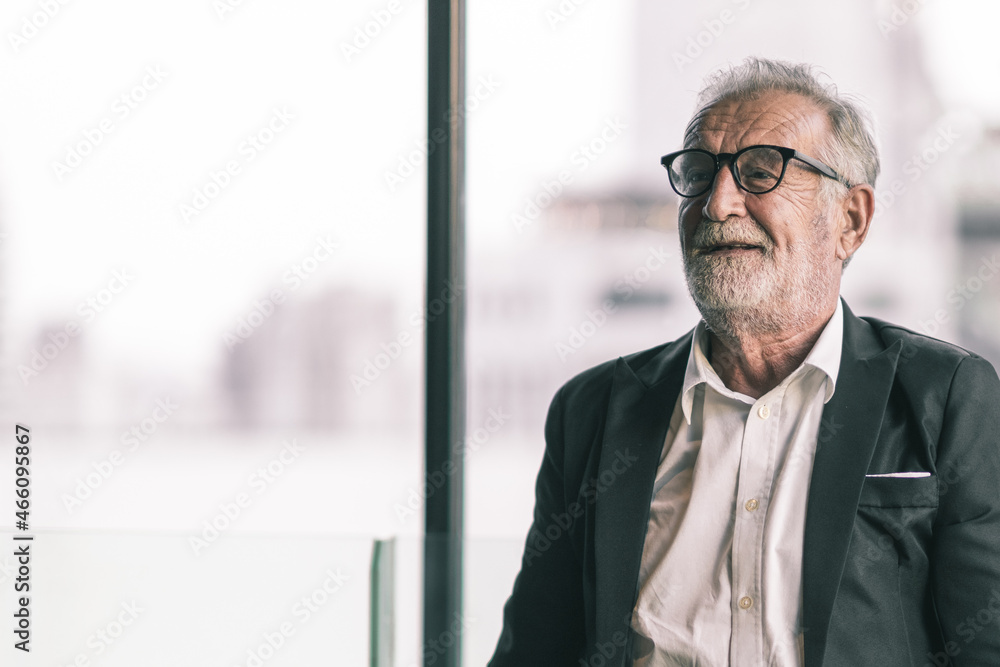 Old business man portrait. He is lookin into the camera with his arm wrap around his body. He is in black suit with white shirt.