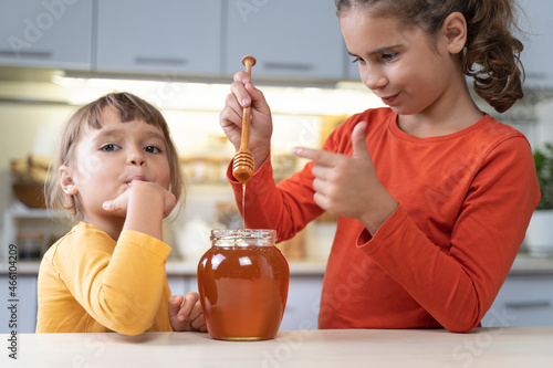 Happy children eating honey from a jar at home in the kitchen. funny girls lick honey from a spoon. Healthy organic food sweets for kids