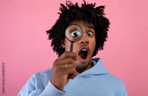 Shocked black teen guy looking through magnifying glass on pink studio background. Secret, doubt concept