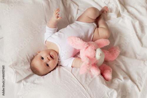 cheerful newborn baby in a white bodysuit lies on a white bed with a plush pink toy rabbit. products for children, toy. concept of a happy childhood and motherhood. child care. space for text.