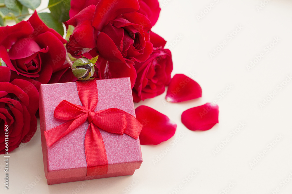 A red gift box for a ring with a red satin ribbon bow on a beige background next to a bouquet of red roses on green stems and red petals with a place for text A red gift box for a ring with a red satin ribbon bow on a beige background next to a bouquet of red roses on green stems and red petals with a place for text