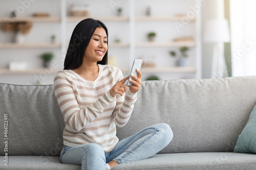 Happy Young Asian Lady Using Modern Smartphone While Resting At Home