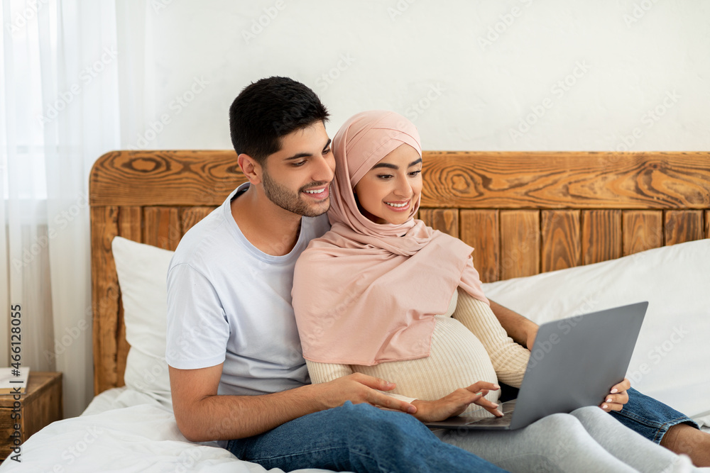Cheerful pregnant arab couple using laptop computer, watching movie together while resting on bed Cheerful pregnant arab couple using laptop computer, watching movie together while resting on bed