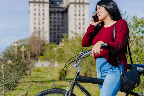 Fototapeta Naklejka Na Ścianę i Meble -  smiling young latina woman talking on her smart phone on a call in a city park with her bicycle next to her