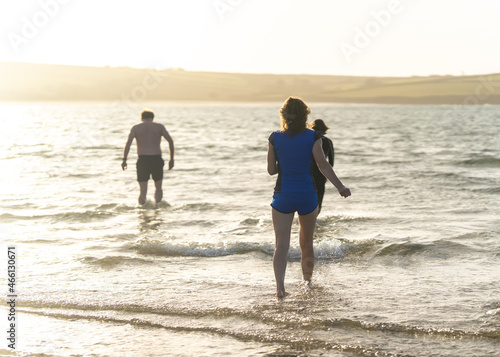 Anonymous man and woman running into freezing cold ocean sea with waves at sunset for natural swimming on sunny afternoon. Beautiful golden hour light in shorts exploring nature with wanderlust.