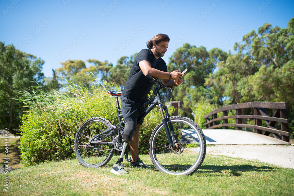 Obraz premium Man with mechanical leg holding mobile phone. Sporty person with disability sitting on bicycle, phone in hands. Sport, disability concept